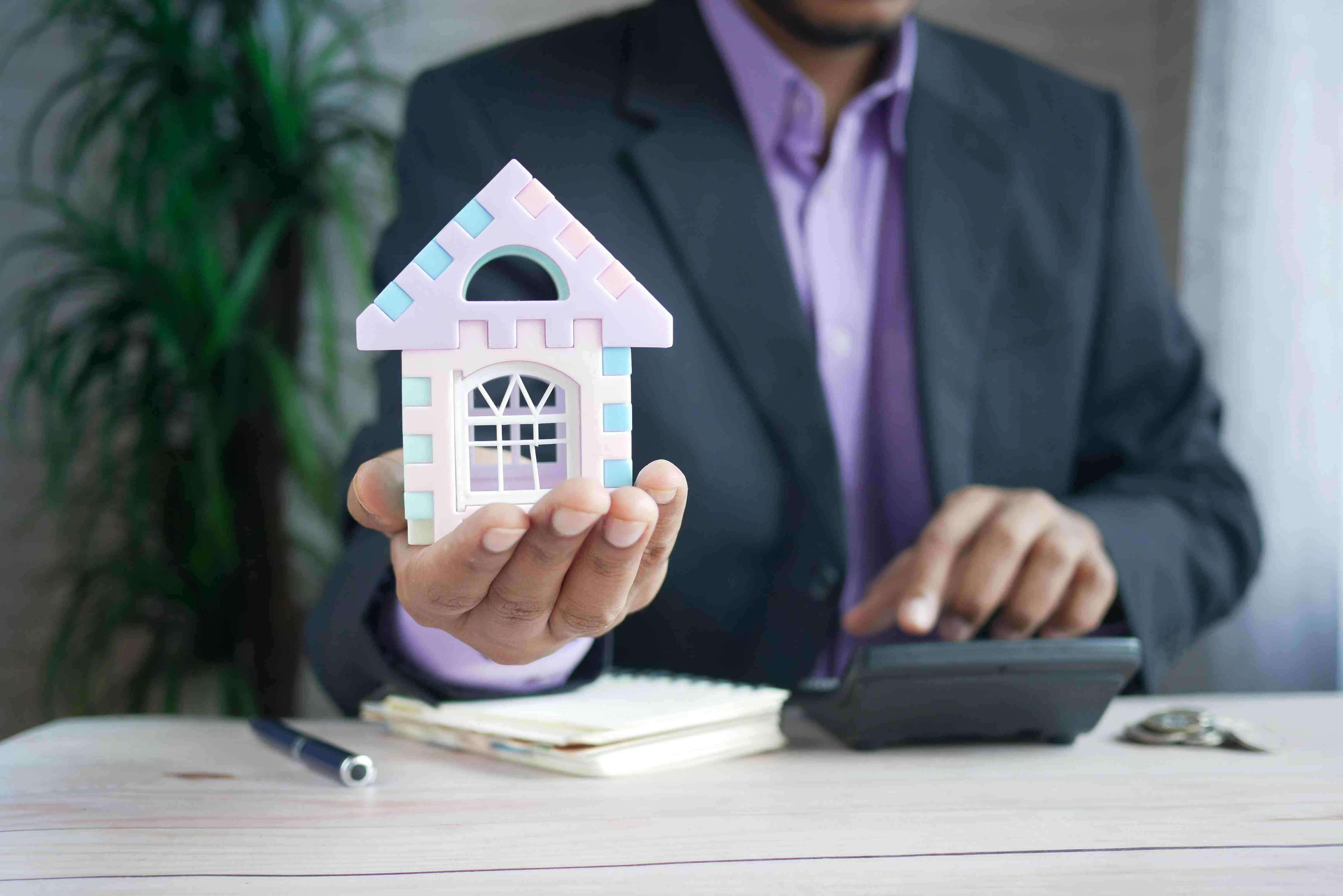 Businessman in a suit holding a small toy house model while using a calculator