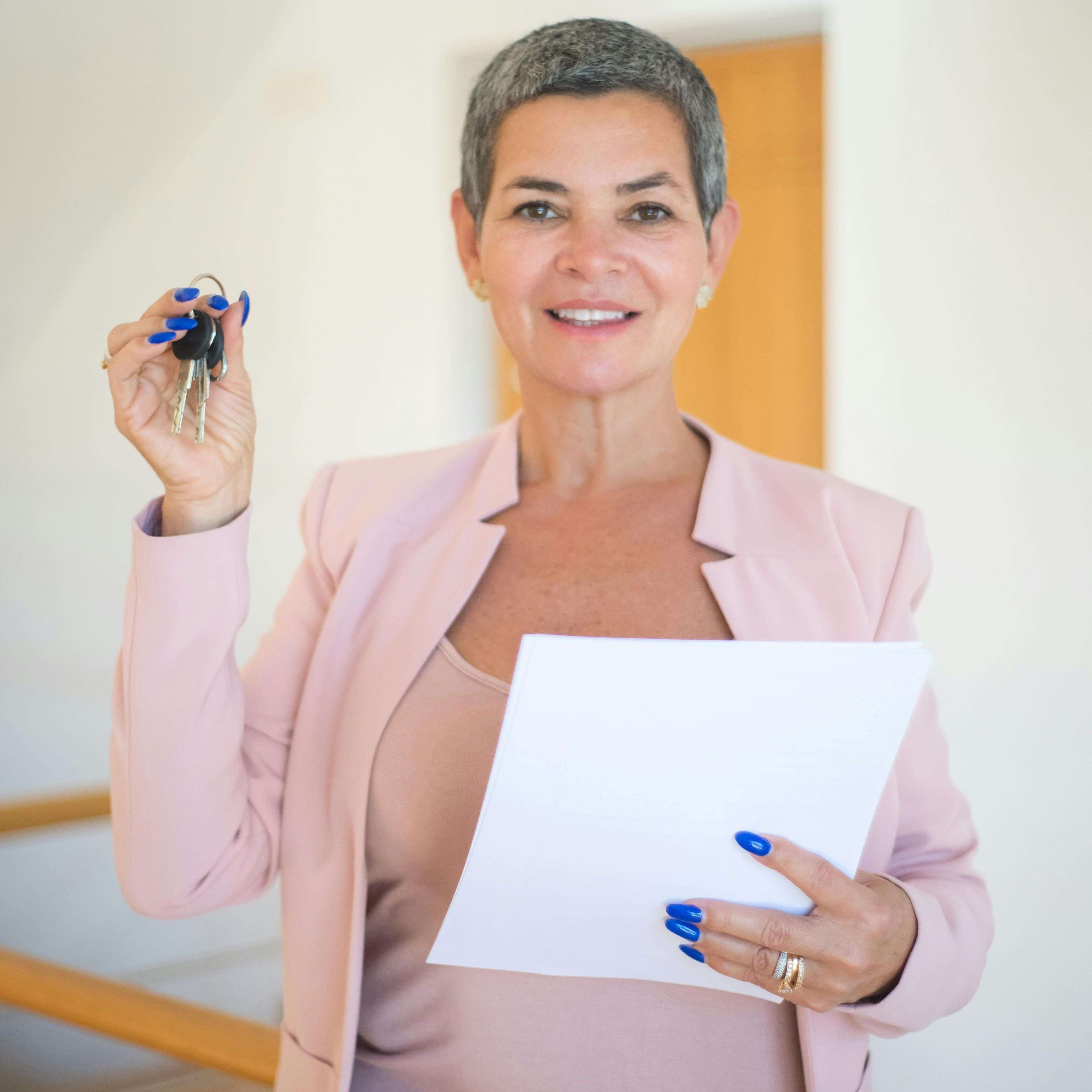 Smiling woman in a pink blazer holds keys and documents indoors, representing real estate success.