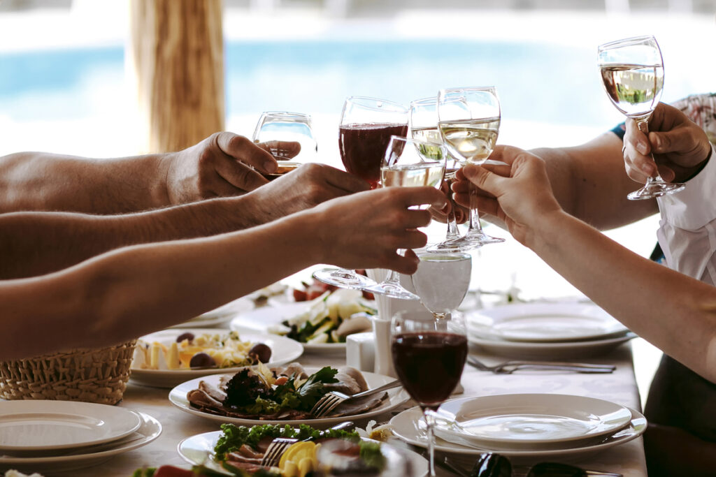 Group of people clinking wine glasses over a shared meal, celebrating together at a table near the coast.