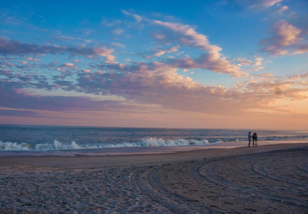 Sunset over a sandy Gulf Coast beach with gentle waves and a few people walking along the shoreline.