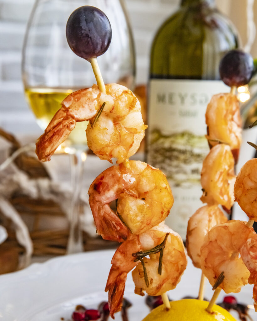 Grilled shrimp skewers garnished with herbs and grapes, served with white wine in a coastal dining setting.