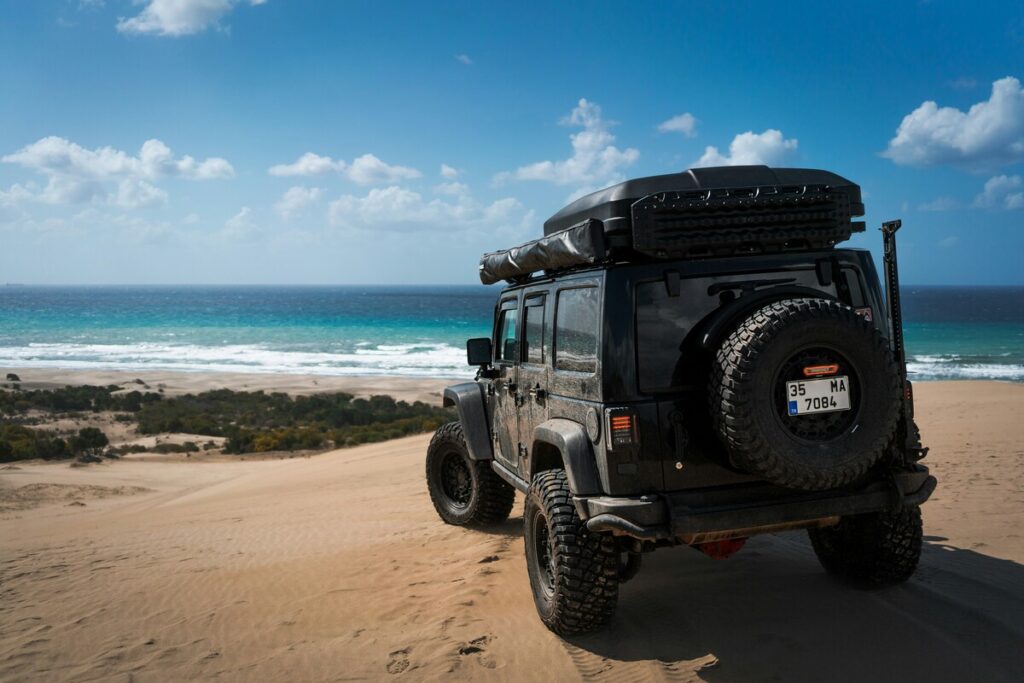 Off-road vehicle parked on sandy dunes overlooking the ocean under a bright blue sky.