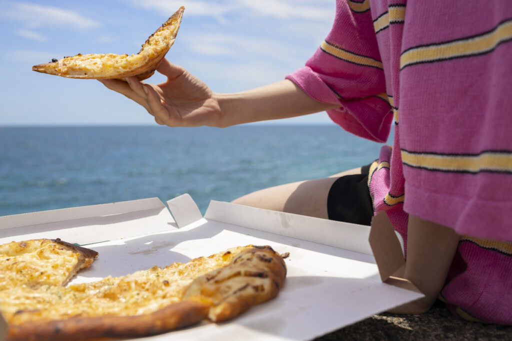 Person holding a slice of pizza from an open box while sitting near the ocean with blue water in the background.