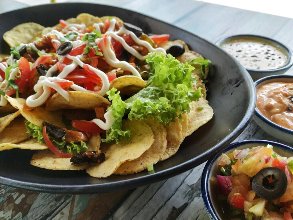 Plate of Tex-Mex nachos topped with tortilla chips, lettuce, olives, tomatoes, and drizzled sauce, served with salsa and dipping sauces at a casual coastal restaurant near San Luis Beach in Galveston, Texas.
