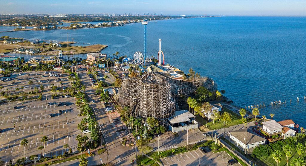 Aerial view of Kemah Boardwalk amusement park on Galveston Bay featuring a wooden roller coaster, Ferris wheel, drop tower, large parking lots, and waterfront attractions